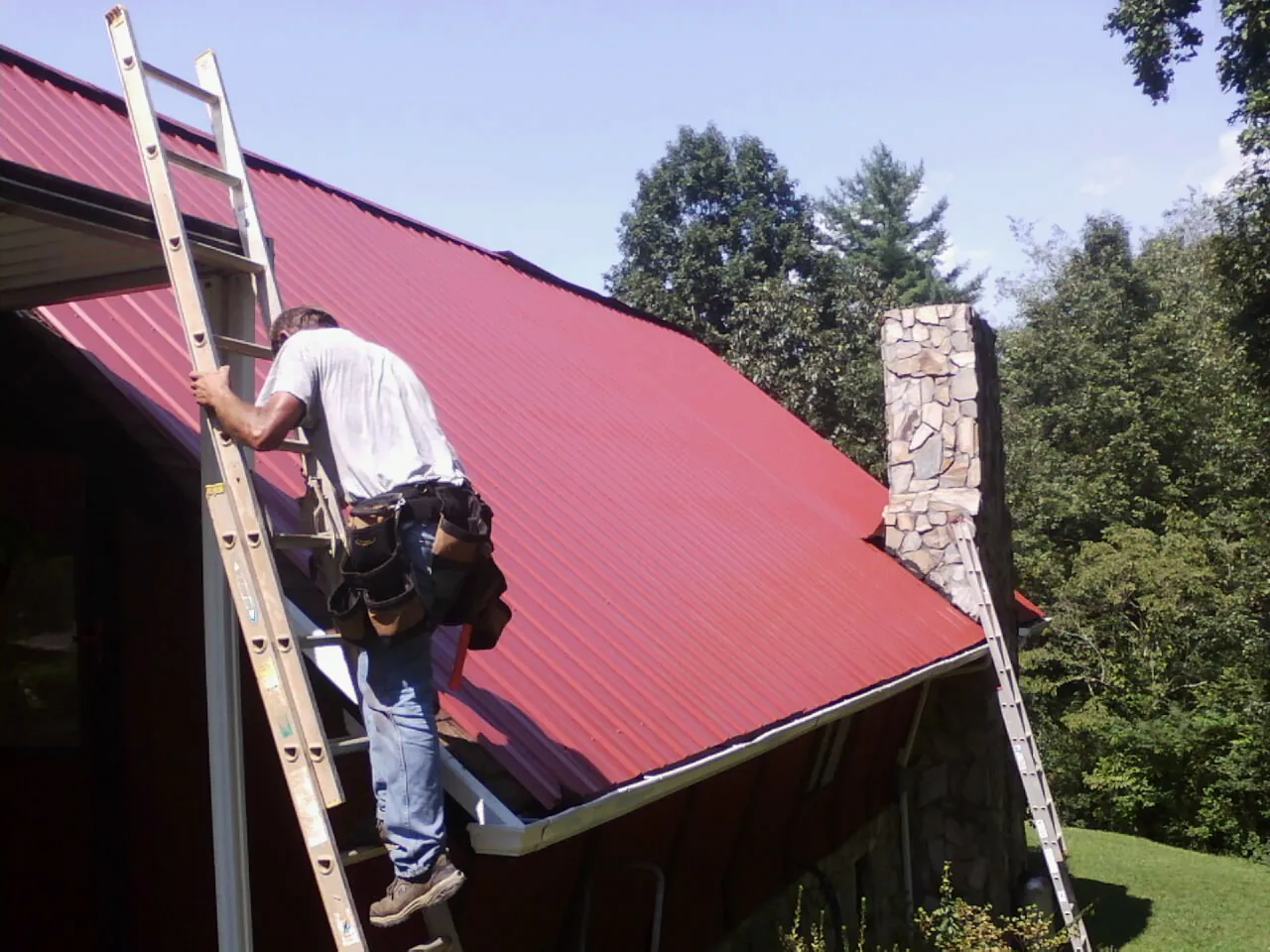 Roofer on a ladder beside a red metal roof and stone chimney — Soffit & Fascia Repair by Roofing Brothers in West Terre Haute
